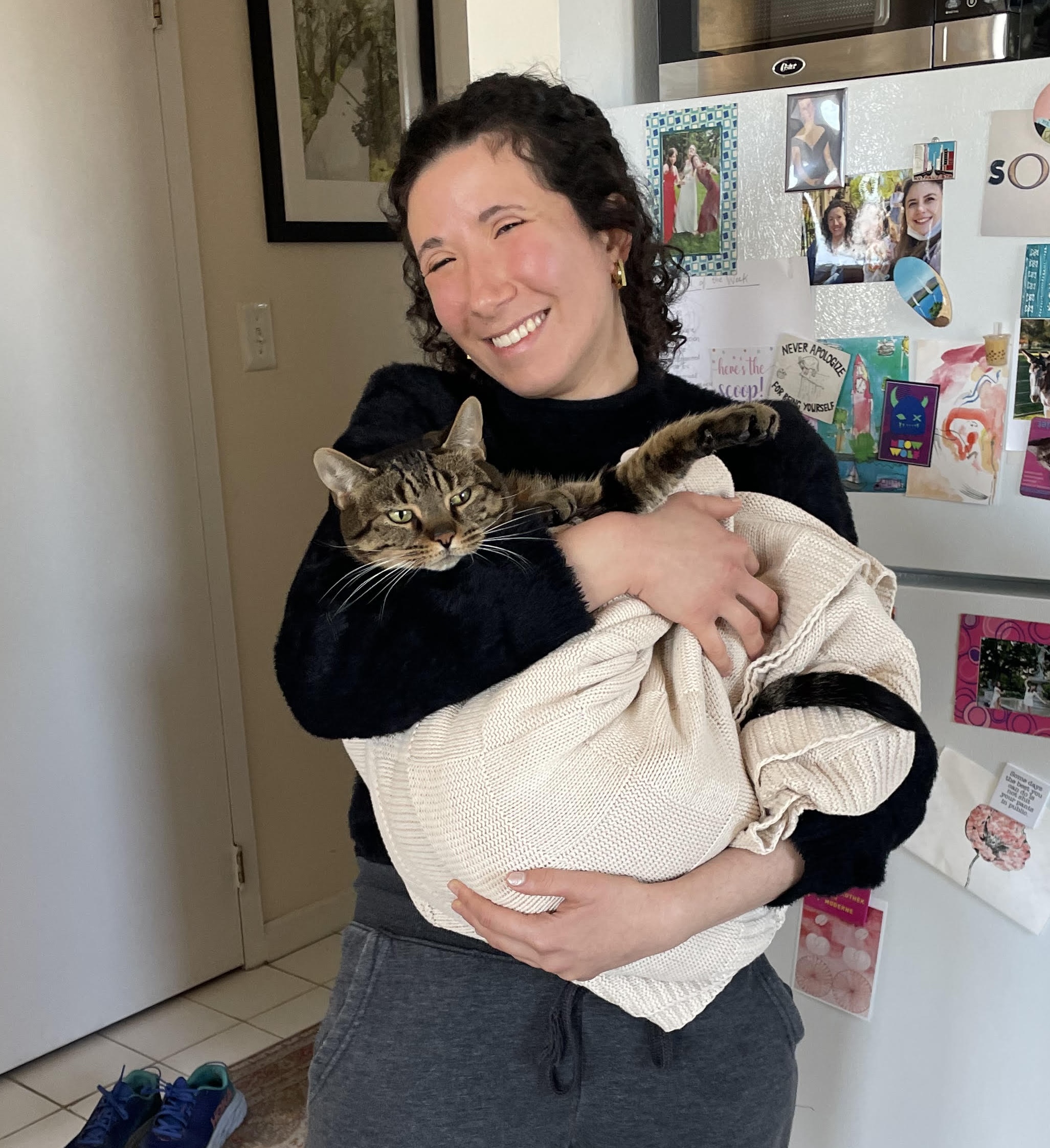 A woman standing in front of a fridge, smiling and holding a cat wrapped in a blanket. The cat looks relaxed and comfy in the blanket. The girl is wearing a black sweater and gray sweatpants. She has curly black hair in a ponytail.