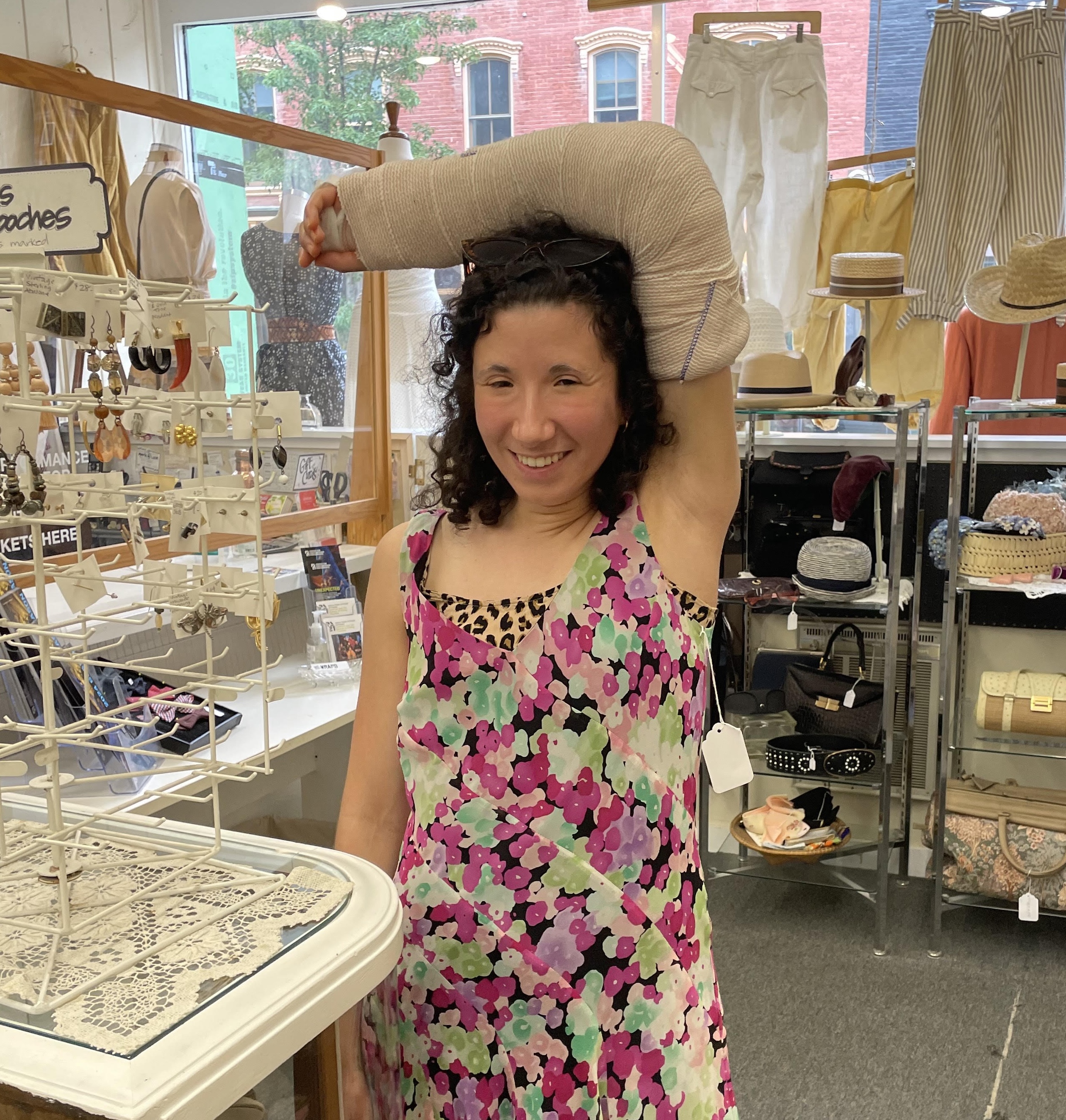 White woman with curly hair wearing a floral dress with her left arm in a giant cast balanced on her head. She is standing in a shop surrounded by knick-knacks and jewelry.